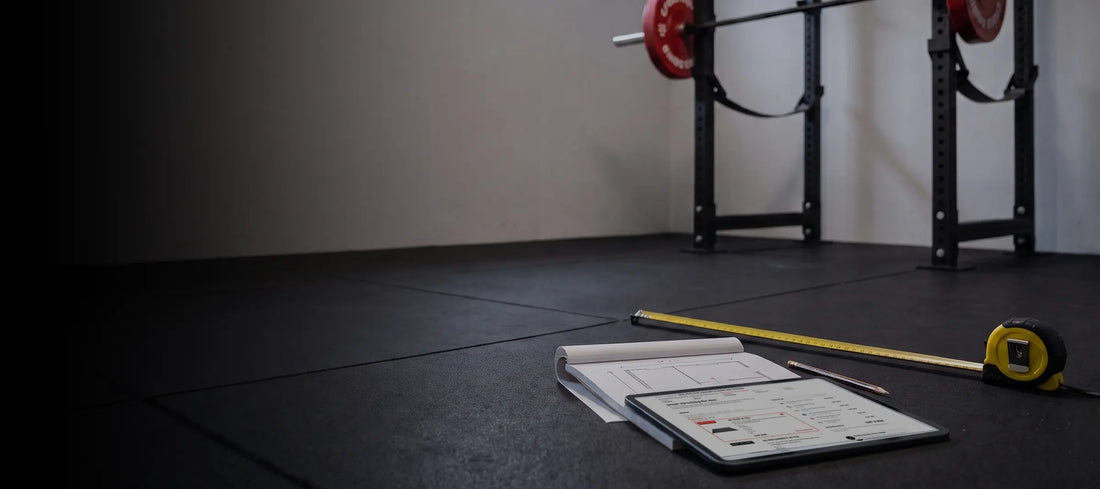 Tablet mit dem Home-Gym-Konfigurator auf einem Notizblock mit Grundriss, daneben Massband und Bleistift auf dem Gummiboden vor einem Power Rack