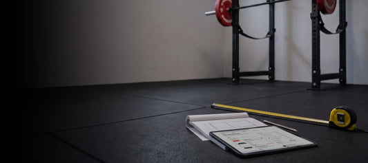 Tablet mit dem Home-Gym-Konfigurator auf einem Notizblock mit Grundriss, daneben Massband und Bleistift auf dem Gummiboden vor einem Power Rack