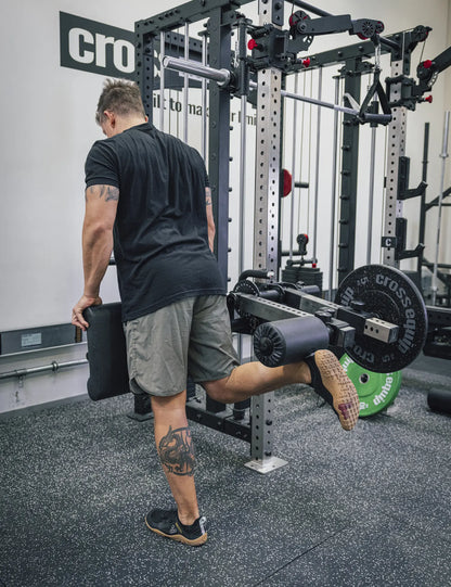 Leg Curl im Stehen am Attachment – eine Hand am Polster, die Rolle hinter dem Unterschenkel, das Knie gebeugt gegen die Last