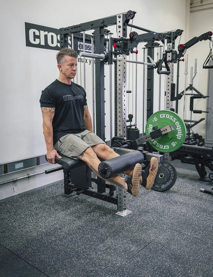 Leg Extension sitzend auf dem Armpolster des Attachments – die Schienbeine drücken die Schaumstoffrolle nach oben, beladen mit einer Bumper Plate