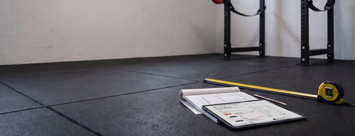 Tablet mit dem Home-Gym-Konfigurator auf einem Notizblock mit Grundriss, daneben Massband auf dem Gummiboden vor einem Power Rack